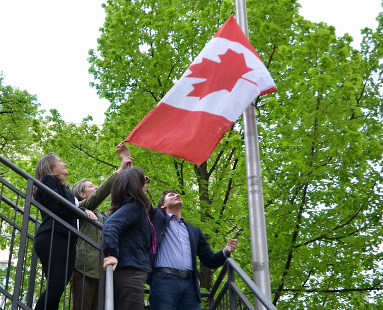 Four people raise the Canadian flag against a backdrop of bright green trees.