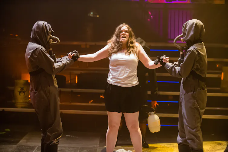 Actors on a stage: In the centre, there appears to be a cis-gendered white woman in a tank top and shorts seen with her hands restrained on both sides by actors wearing gas mask type equipment.