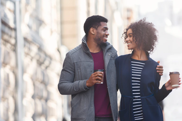 A young couple walks arm in arm, holding coffee cups and smiling at each other