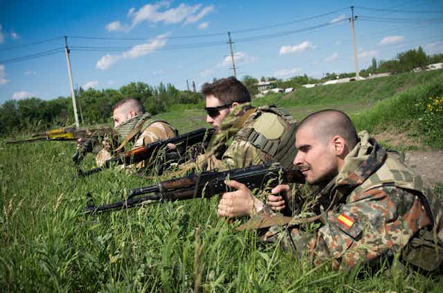 Three people in camouflage clothes lie on the ground pointing rifles