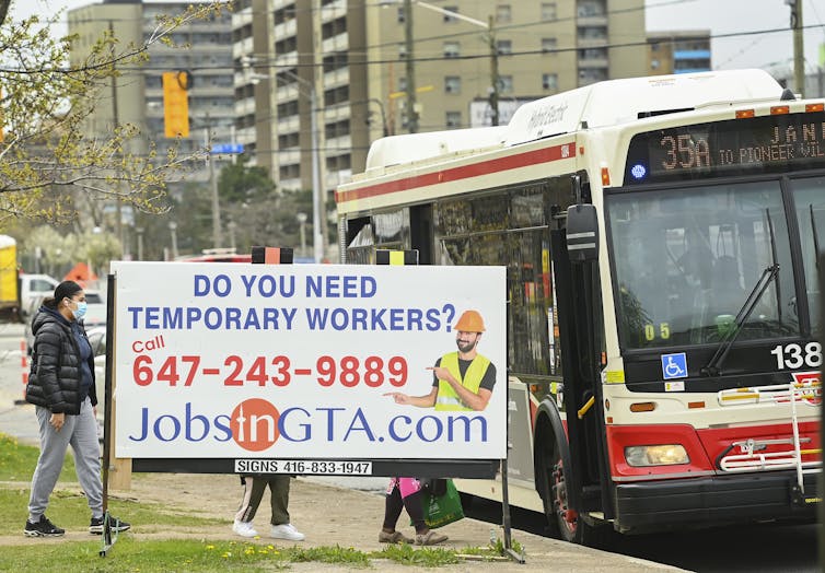 People are seen walking behind a sign for temporary jobs while stepping onto a city bus.