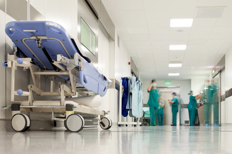 An empty gurney in the foreground of a hospital corridor, with people in scrubs out of focus in the background