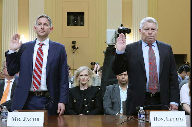 two men in business suits stand at a table with their right hands raised