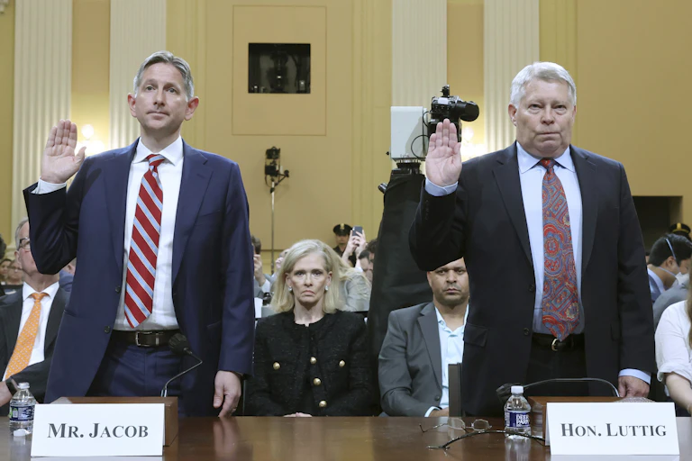 two men in business suits stand at a table with their right hands raised