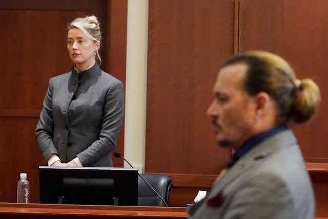 Amber Heard wears a grey suit and stands next to Johnny Depp, seated.