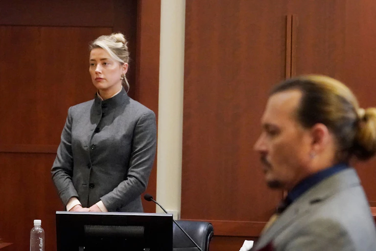 Amber Heard wears a grey suit and stands next to Johnny Depp, seated.