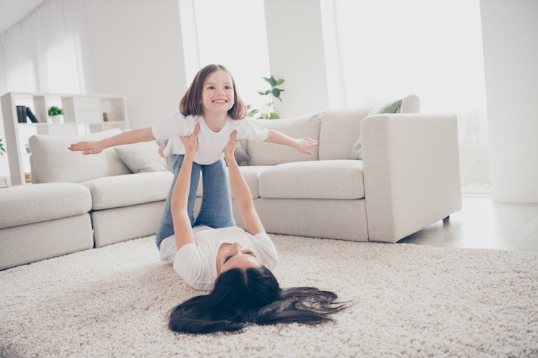 A woman lying on her back on a white carpet holds up a little girl who is pretending to fly.  There is a white couch behind them.