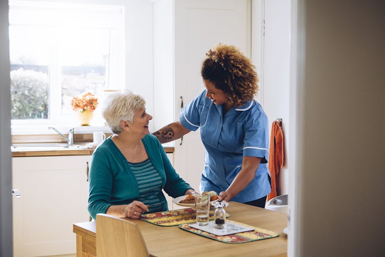 A carer gives an elderly lady her dinner in a kitchen setting.