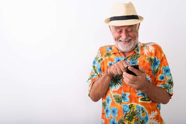 An older white man in a Hawaiian shirt and panama hat laughing at his phone.