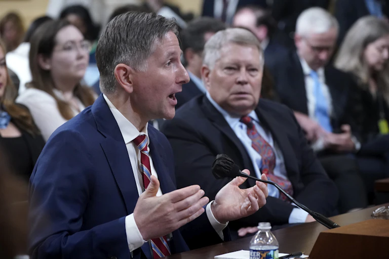 Two men, both wearing dark blazers and ties, sit at a table in a crowded room; one talks into a microphone.
