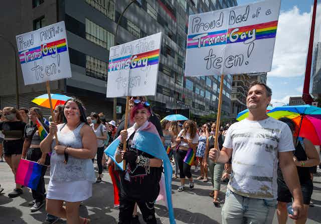 Parents and a trans teen seen at a pride parade with signs saying 'proud mom of a trans gay teen' and 'proud dad of a trans gay teen.' 