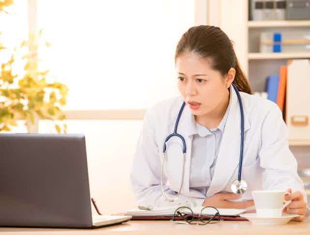 Woman wearing a white coat with a stethoscope seated at a desk looking at a laptop with a look of disbelief