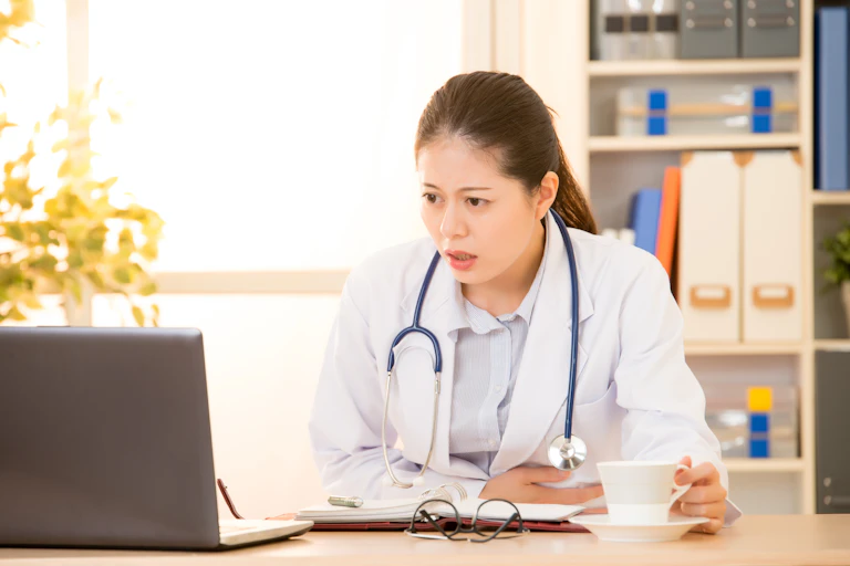 Woman wearing a white coat with a stethoscope seated at a desk looking at a laptop with a look of disbelief