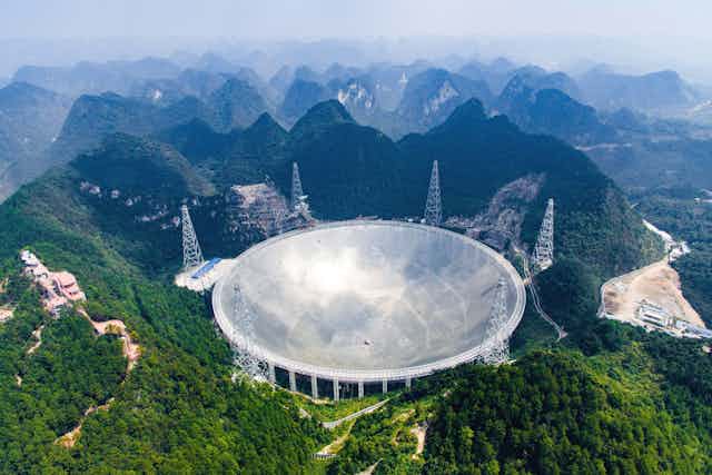 Fotografía aérea que muestra una enorme antena parabólica de metal plateado enclavada entre verdes colinas montañosas.