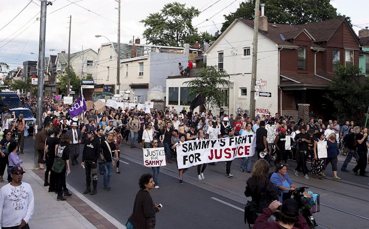 a crowd of people protest against police brutality with a sign reading SAMMY'S FIGHT FOR JUSTICE