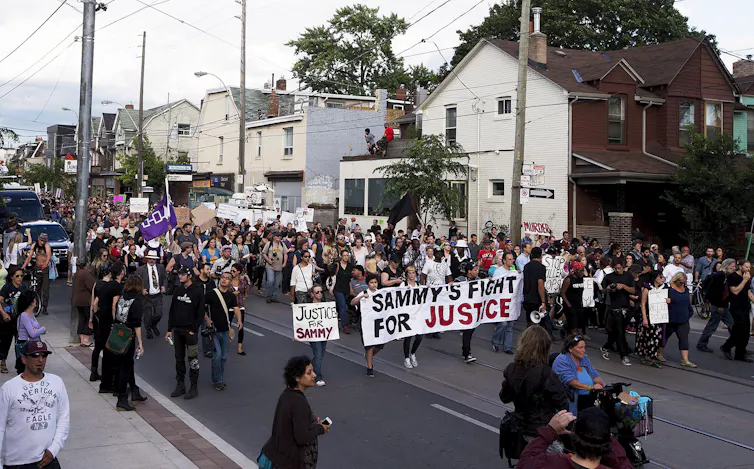a crowd of people protest against police brutality with a sign reading SAMMY'S FIGHT FOR JUSTICE