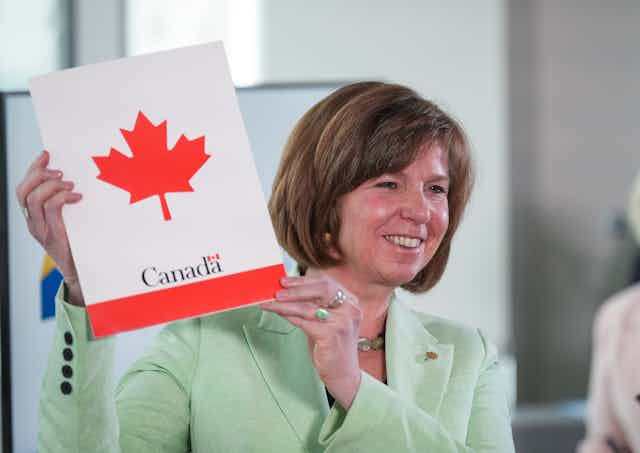 A woman in a green jacket smiling and holding up a document with the word Canada on it and a maple leaf