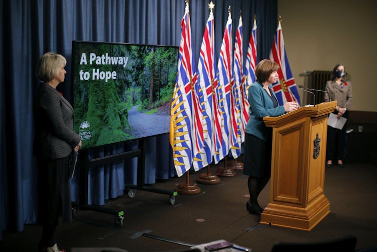 A woman at a podium in front of a row of flags, and two other women at opposite sides of stage, in front of background with the words 'A pathway to hope'