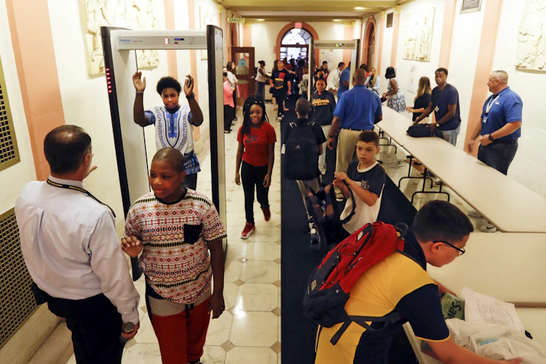 Students walk past tables with adults opening bags, and through a metal detector