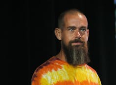 A bald man with a thick graying beard and a bright yellow and orange tie-dye shirt looks off into the distance.