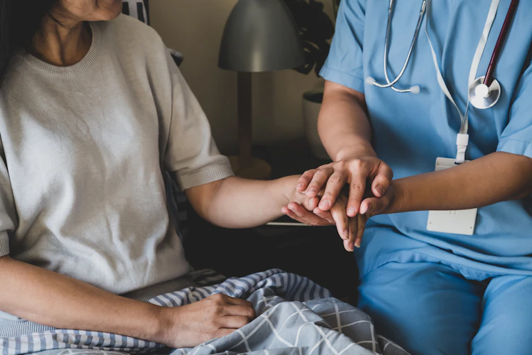A woman in a white shirt sits in bed beside a doctor wearing a stethoscope who holds her hand.