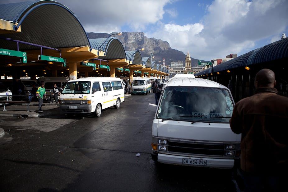 Two orderly queues of white minibus taxis are parked alongside signs denoting various destinations. Table Mountain can be seen in the background.