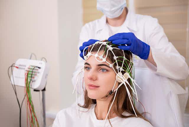 Young woman with nodes and wires on her head for an EEG brain scan, while a doctor with blue gloves adjusts