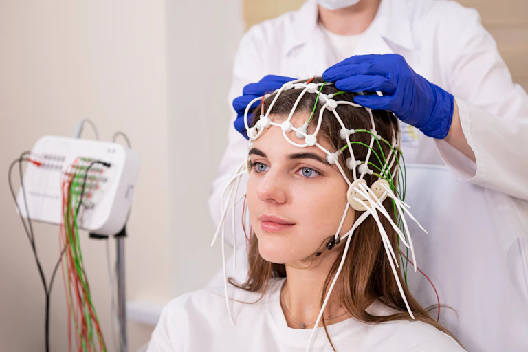 Young woman with nodes and wires on her head for an EEG brain scan, while a doctor with blue gloves adjusts