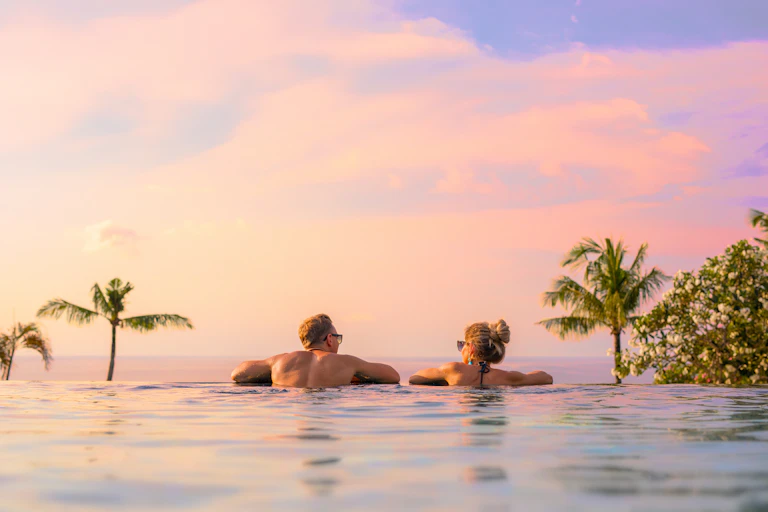 Two people immersed in water look out over a sunset sky with trees on the left and right