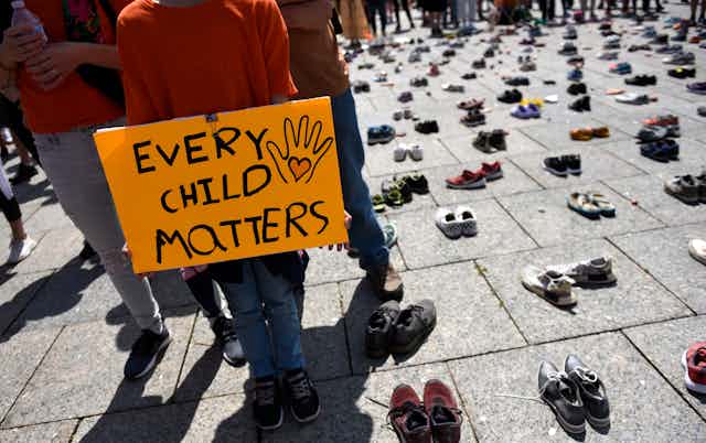 Dozens of pairs of children's shoes on the pavement. A person in an orange T-shirt stands beside the shoes with a sign that reads Every Child Matters.
