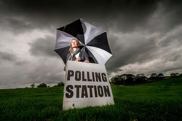 A woman sits in a field under an umbrella holding a sign directing people to a polling station. 