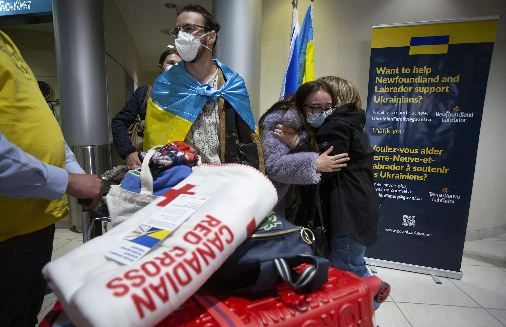 A man is wrapped in a Ukrainian flag behind a luggage cart as a woman hugs another woman behind him