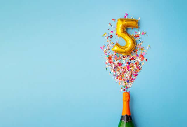 A gold balloon shaped like the number five is surrounded by confetti that appears to be shooting out of a champagne bottle.