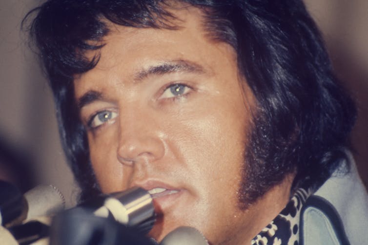 Man with blue eyes and sideburns speaks into microphone.