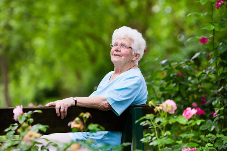 An elderly woman sitting on a bench in the garden looking happy.