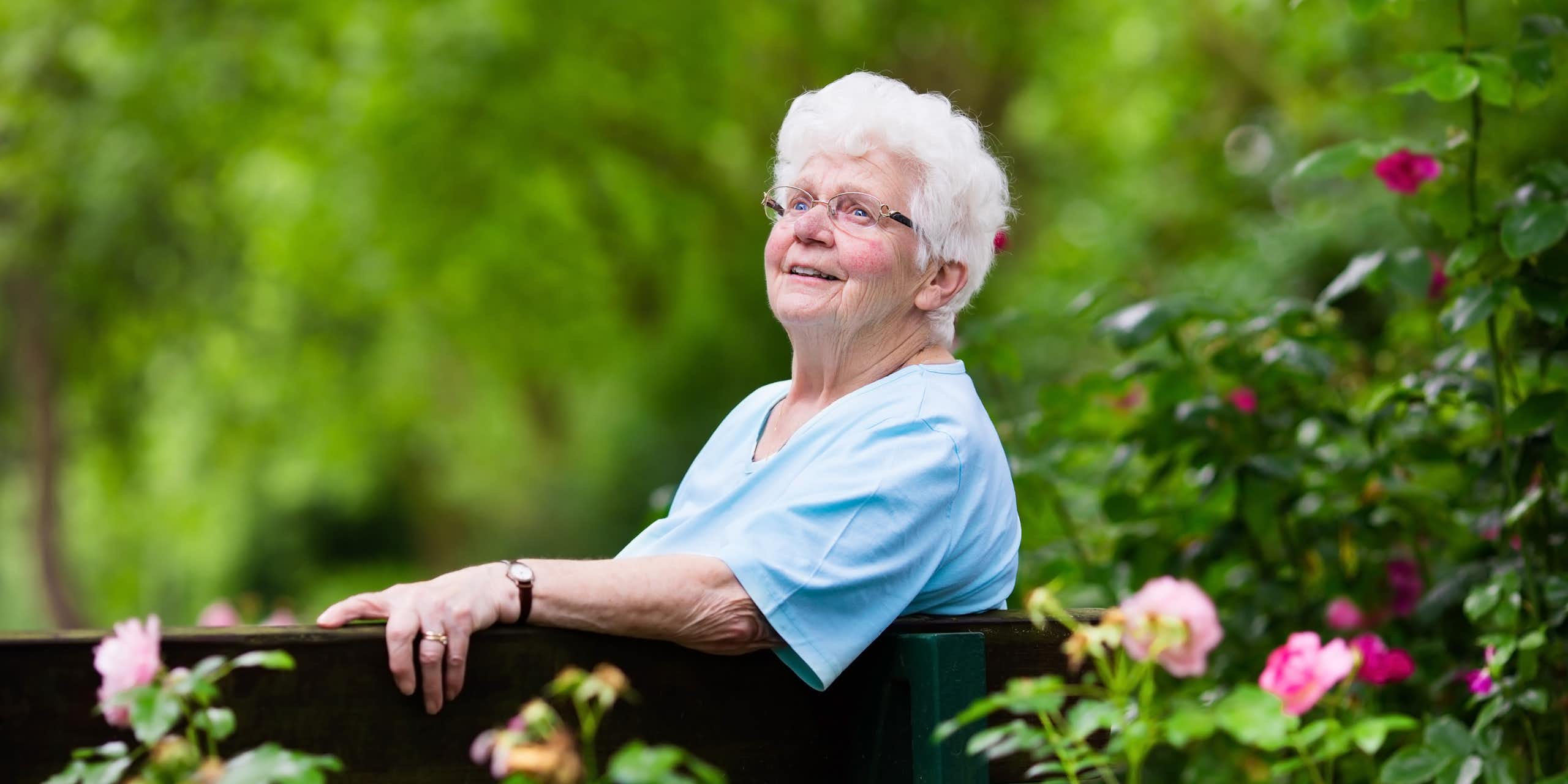 An elderly woman sitting on a bench in the garden looking happy.