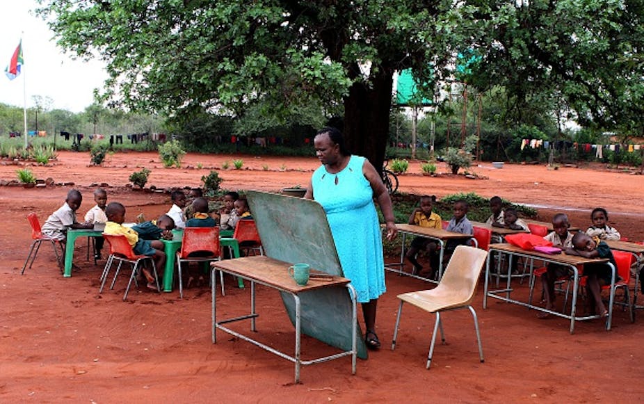 Pupils sitting under a tree watch as a female teacher writes on a small chalkboard balanced against a school desk.