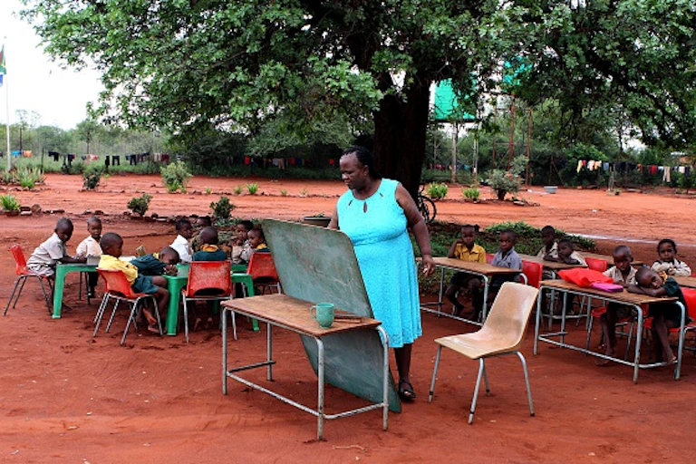 Pupils sitting under a tree watch as a female teacher writes on a small chalkboard balanced against a school desk.