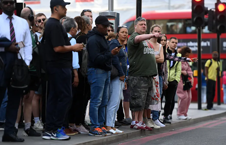 Large group of people queuing in the street, red double-decker bus in the background