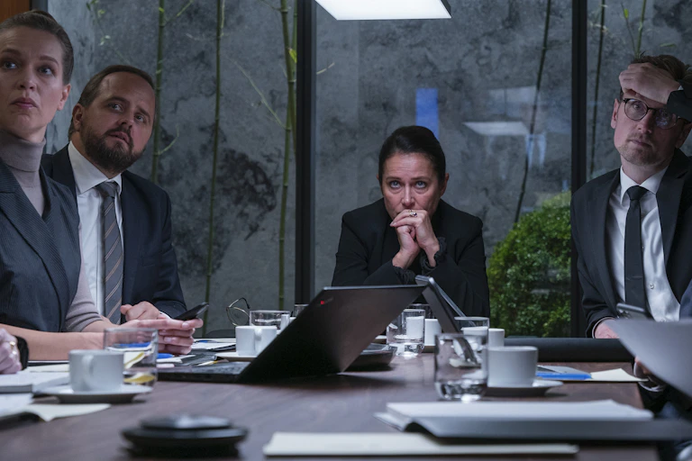 A scene from Borgen showing Nyborg at the head of a table, surrounded by advisers, looking concerned with her hands clasped in front of her mouth