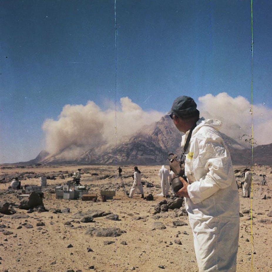 Des hommes en tenue de protection regardent une explosion dans un désert.