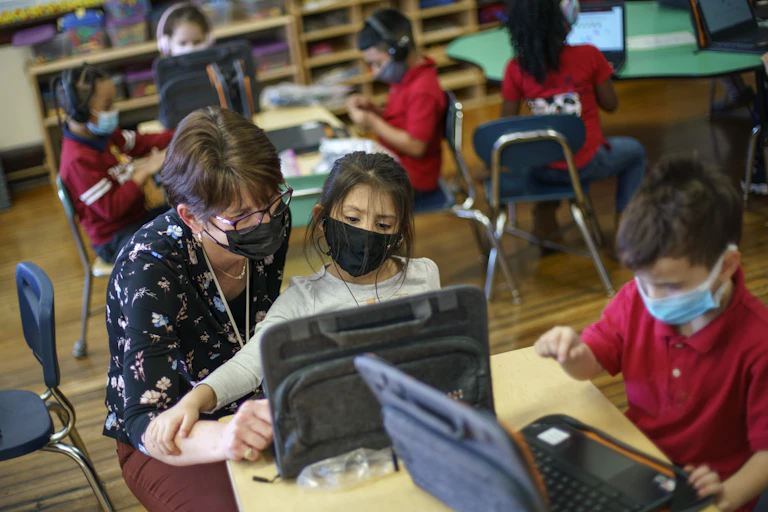 A teacher crouches next to a young student at a desk