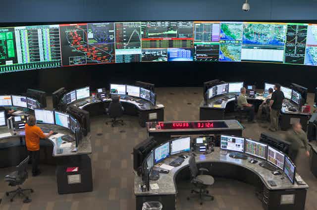 A large room with a wall of electronic displays and managers seated at computers