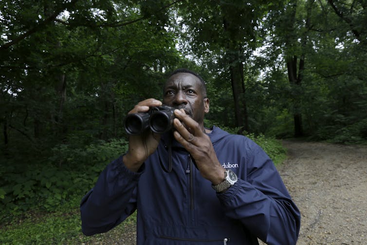 A black man holding binoculars in a forest.