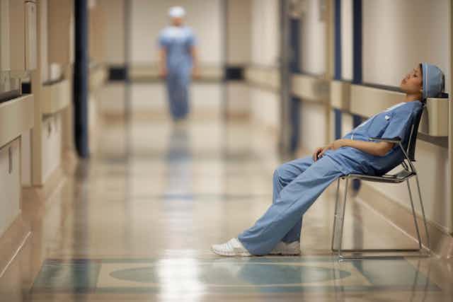 Female medical professional in blue scrubs sitting in a chair and leaning against the wall in a hallway resting.