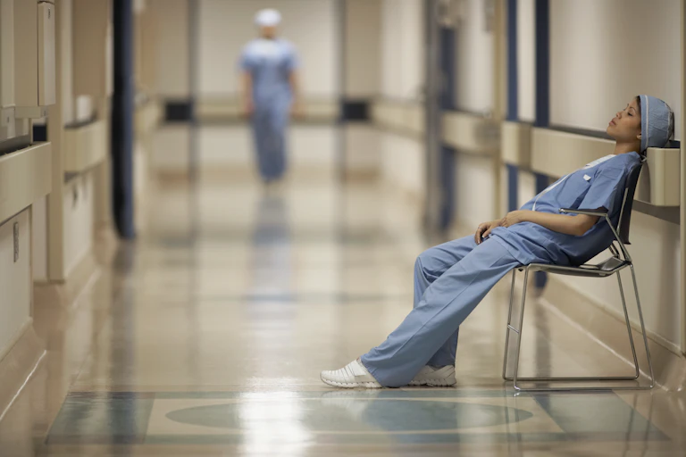 Female medical professional in blue scrubs sitting in a chair and leaning against the wall in a hallway resting.