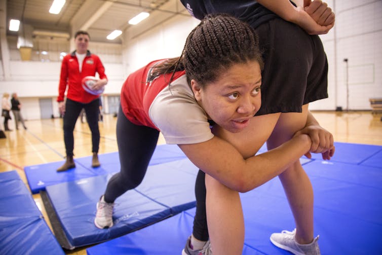 a young woman tackles another standing woman in a gym as a coach watches on