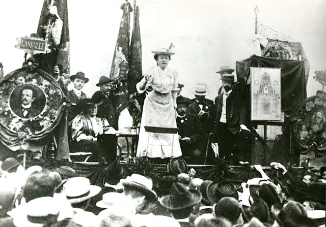 A woman in a white long dress and hat giving a speech from an outdoor stage surrounded by people.