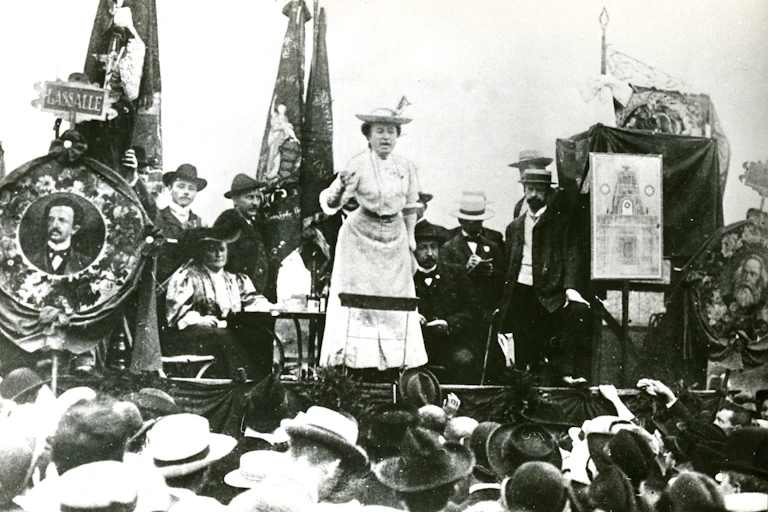 A woman in a white long dress and hat giving a speech from an outdoor stage surrounded by people.