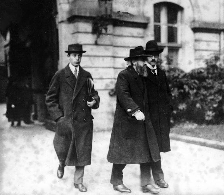 Tres hombres con abrigos y sombreros de copa se alejan de un gran edificio.